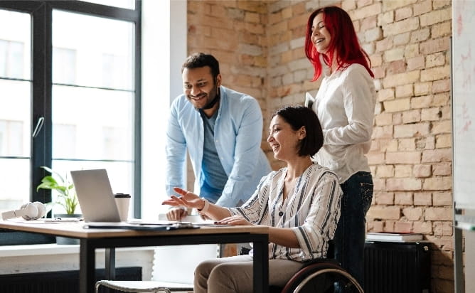 a disabled woman in a wheelchair sitting at a desk showing her coworkers something